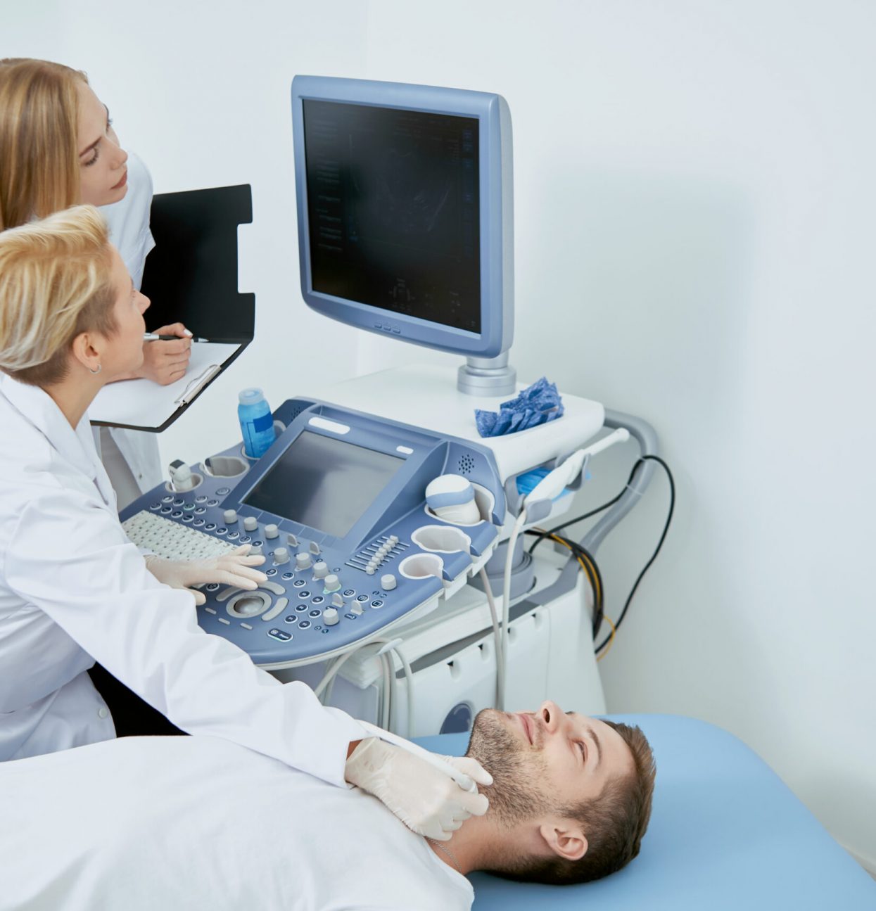 Side view of three people on medical consultation. Pofessional doctor sitting working examining patient looking at monitor and young assistant standing near and helping her.